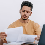 A person reviewing paperwork at a desk beside a laptop.